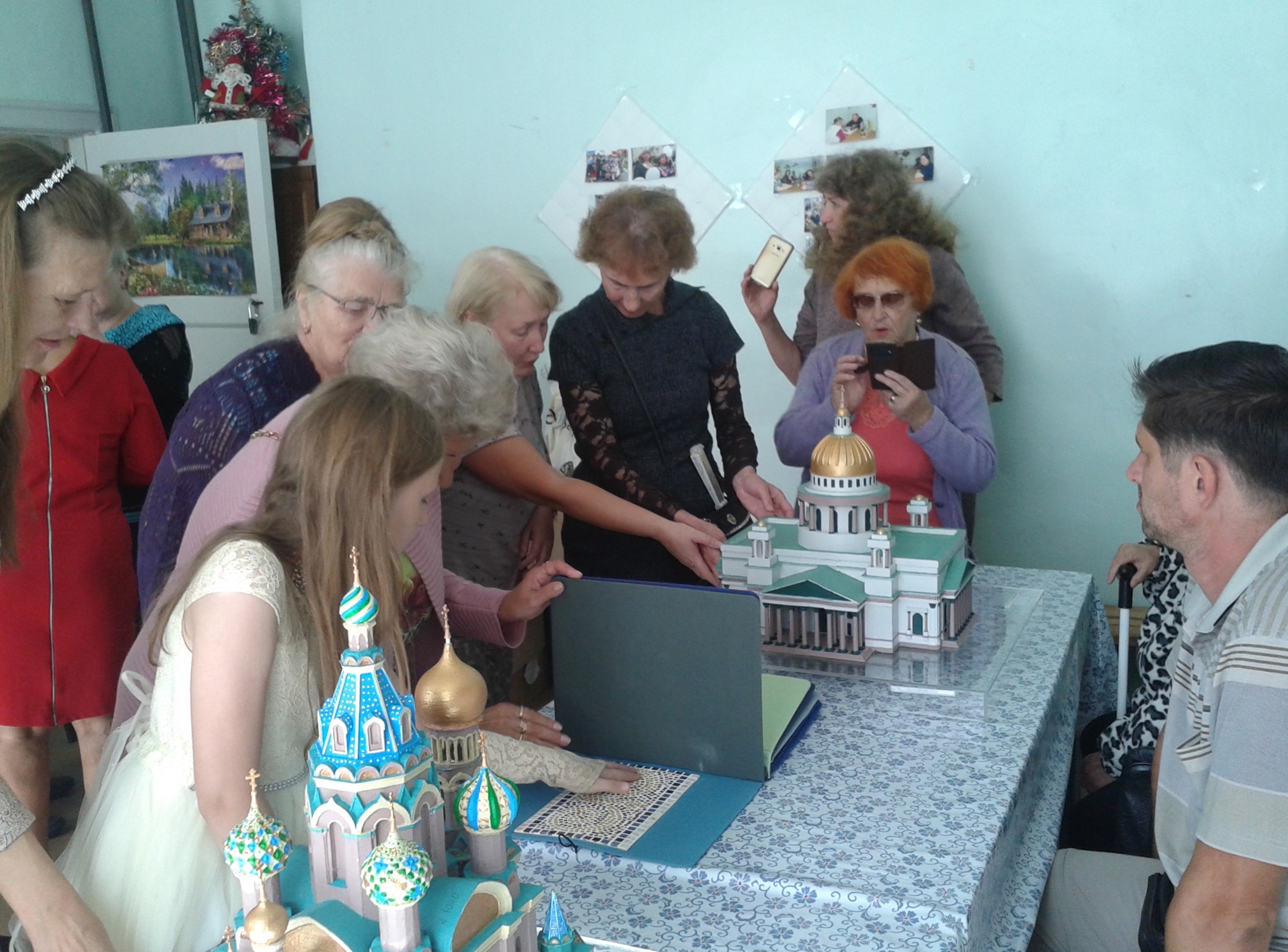 На горизонтальной фотографии – мероприятие в Сланцевской местной организации ВОС. На переднем плане на столе стоят 3D-модели и тактильная книга, которые тактильно изучают посетители, стоящие за столом лицом к камере.  