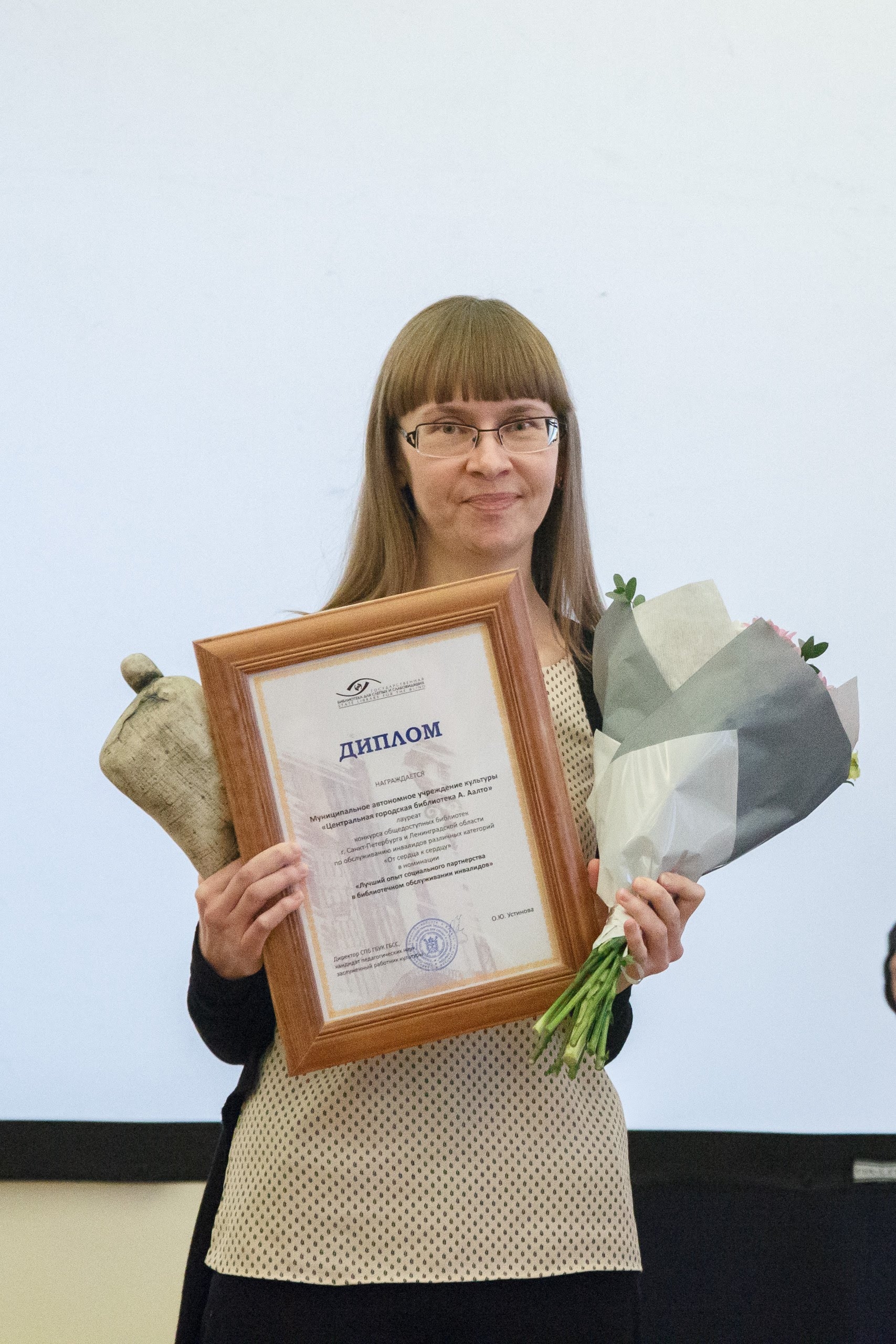 На вертикальной фотографии на переднем плане стоит женщина, которая держит в правой руке статуэтку и диплом победителя, в левой руке - букет цветов. На заднем плане расположен белый экран.