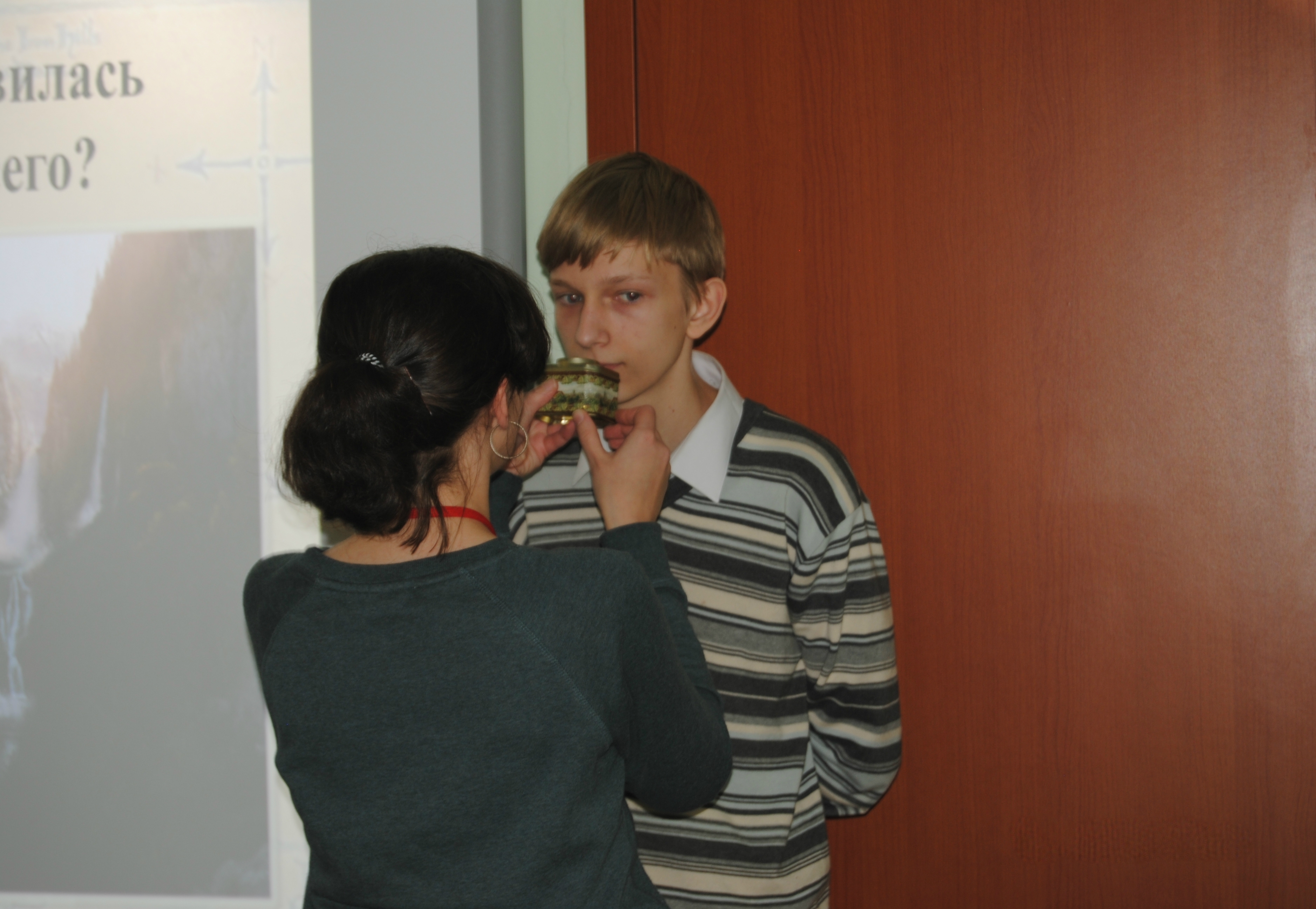 На горизонтальной фотографии изображен подросток, который пытается определить запах из ёмкости. Ёмкость держит ведущая мероприятия