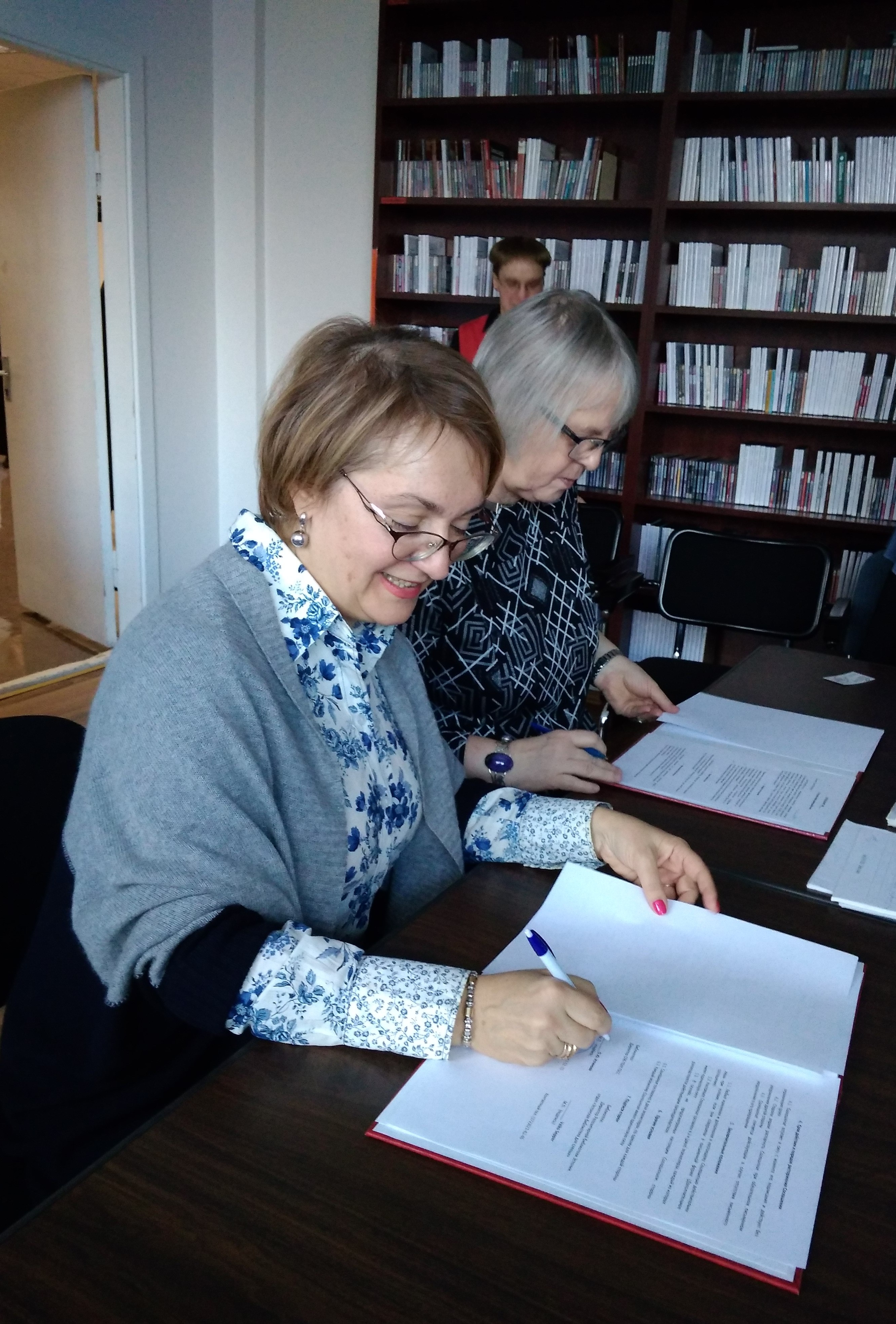 На вертикальной фотографии на переднем плане за столом сидят две женщины, которые подписывают документы. 