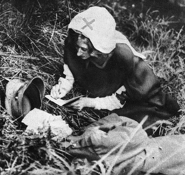 Черно-белая вертикальная фотография. Сестра милосердия в белом платке записывает в блокнот слова лежащего рядом раненого солдата.