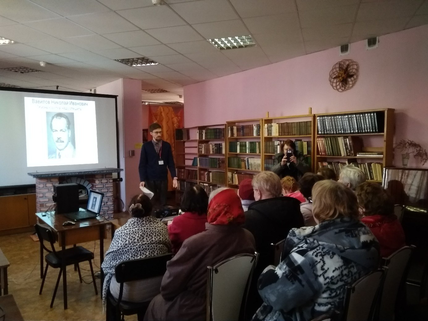 На горизонтальной фотографии на переднем плане в несколько рядов спиной к камере сидят зрители пенсионного возраста. Перед ними стоит сотрудник Библиотеки.