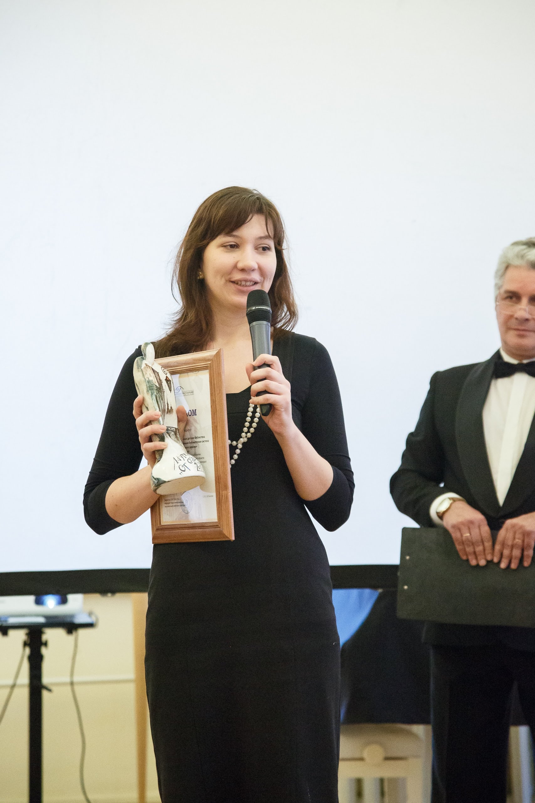 На вертикальной фотографии на переднем плане стоит женщина, которая держит в правой руке статуэтку и диплом победителя, в левой руке - микрофон. На заднем плане расположен белый экран.