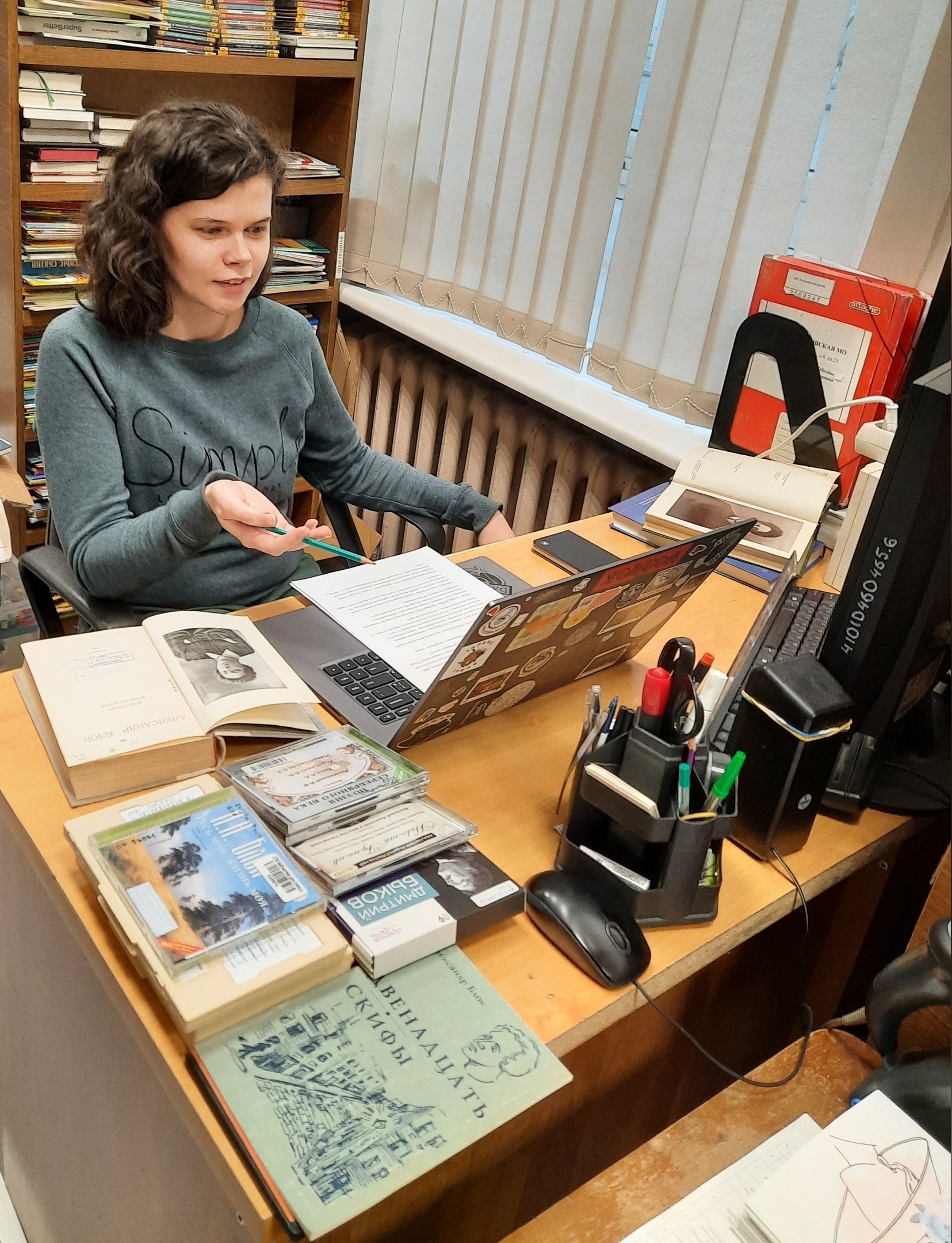 Лицом камере за столом, на котором лежат ноутбук и книги, ведущая рассказывает о Блоке