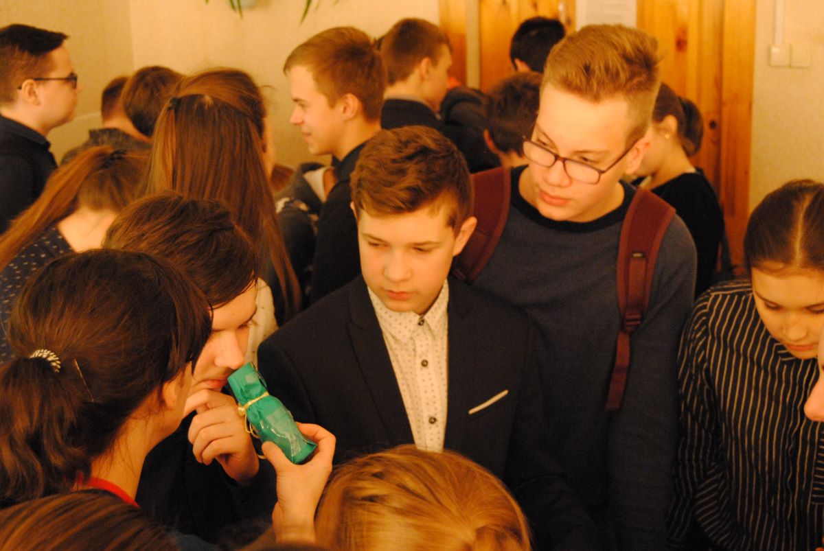 На горизонтальной фотографии стоят школьники подросткового возраста вместе с ведущей мероприятия. Один из участников пытается определить запах из флакона, который подносит ему ведущая. Взгляды некоторых ребят обращены на флакон.