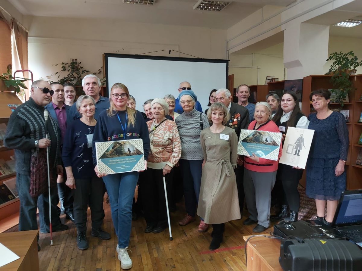 На горизонтальной фотографии изображен итоговый момент встречи. Участники и ведущие мероприятия стоят перед экраном лицом к камере. У некоторых из них в руках многоформатные альбомы и рельефно-графическая иллюстрация памятника Шишкину.